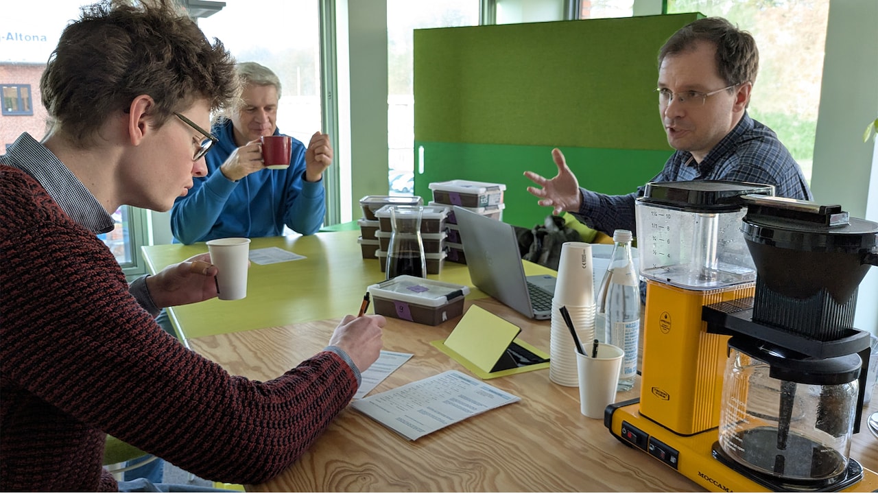 Bei einer Kaffee-Blindverkostung sitzen drei Personen am Tisch. Zwischen ihnen: Laptop, nummerierte Kaffeedosen ohne sonstige Aufschrift, Becher, eine Kaffeemaschine und viele Umfragezettel.
